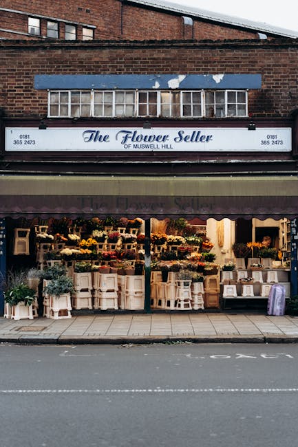 The storefront of 'The Flower Seller' of Muswell Hill, featuring a vibrant display of fresh flowers arranged on multiple white shelves and tables under an awning. The floral arrangements include roses