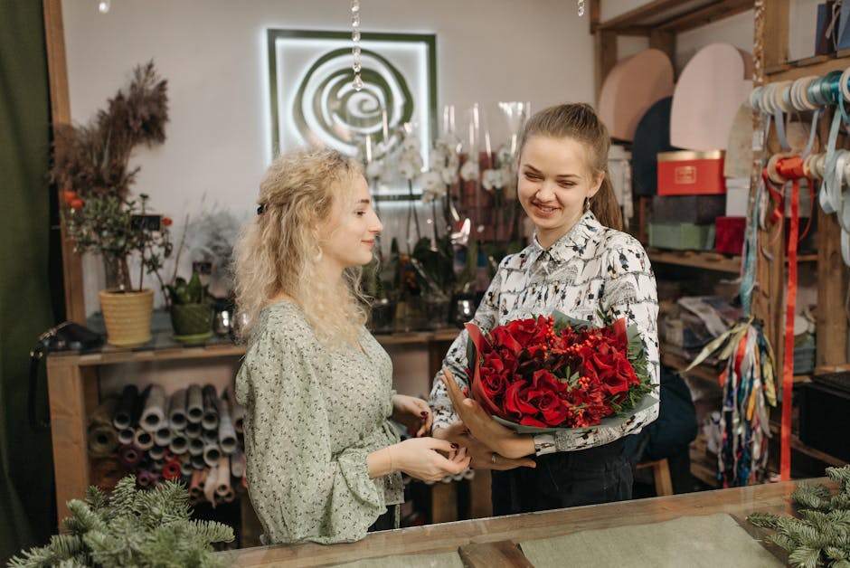 A young woman with light brown hair tied back into a ponytail is smiling as she receives a vibrant bouquet of fresh red roses, red tulips, and green foliage from a florist behind the counter. The flor
